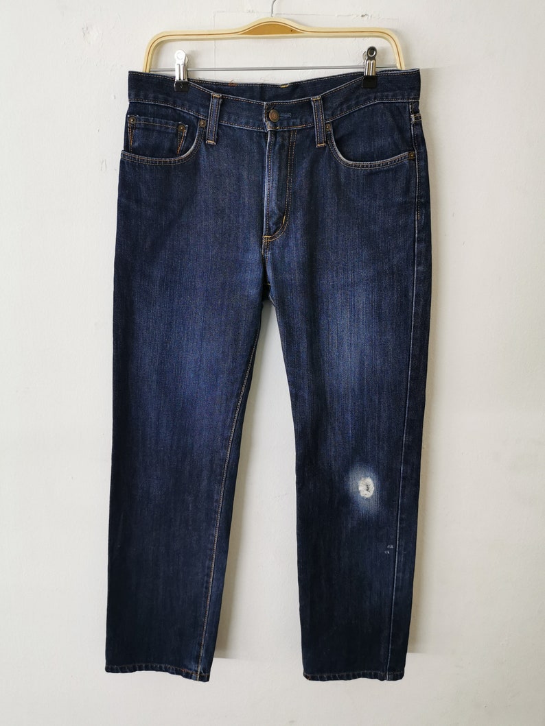 May include: Dark wash denim jeans with a classic five-pocket design. The jeans are hanging on a wooden hanger against a white background. There is a light-colored patch on the right leg.