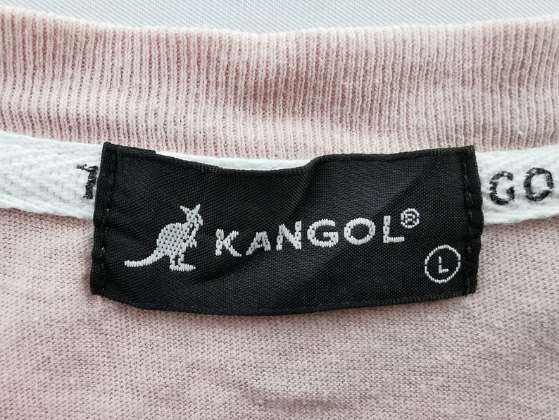 May include: Close-up of a pink t-shirt with a black label. The label features the white text "KANGOL" with a kangaroo logo and the size "L". A white tag with the text "GO" is visible.