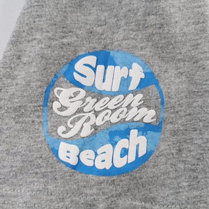 May include: Close-up of a gray t-shirt with a circular graphic. The graphic features the words "Surf Green Room Beach" in white, outlined by a blue circle with a wave design. The shirt is made of a soft, textured fabric.