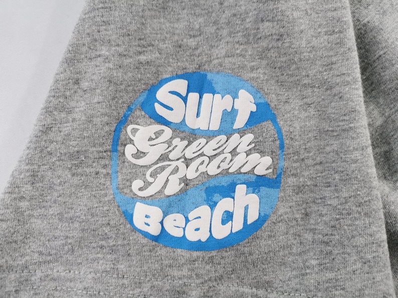 May include: Close-up of a gray t-shirt with a circular graphic. The graphic features the words "Surf Green Room Beach" in white, outlined by a blue circle with a wave design. The shirt is made of a soft, textured fabric.