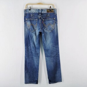 May include: Blue denim jeans with decorative stitching on the back pockets and a small red tag. The jeans are hanging on a wooden hanger against a white background. The jeans have a faded wash.
