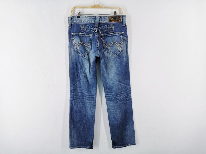 May include: Blue denim jeans with decorative stitching on the back pockets and a small red tag. The jeans are hanging on a wooden hanger against a white background. The jeans have a faded wash.