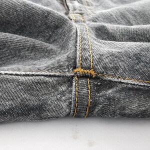 May include: A pair of charcoal gray denim jeans featuring a prominent crotch seam with thick, golden-brown contrast stitching. The fabric displays a classic twill weave texture, and the heavy-duty thread reinforces the structural seams of the garment.