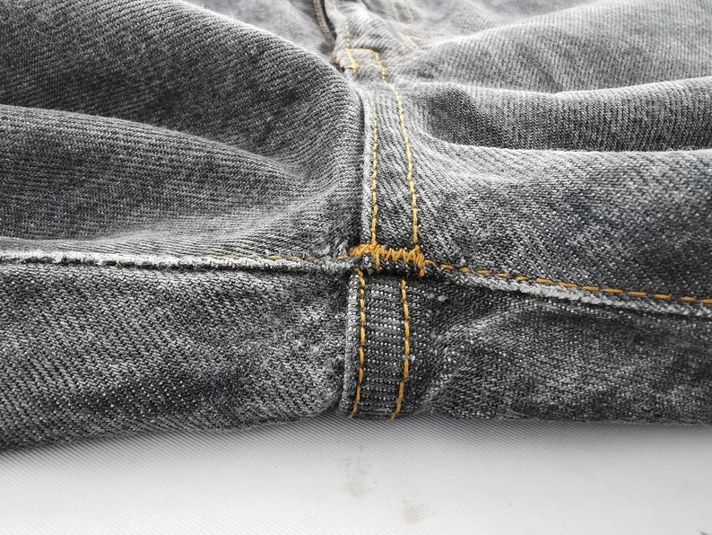 May include: A pair of charcoal gray denim jeans featuring a prominent crotch seam with thick, golden-brown contrast stitching. The fabric displays a classic twill weave texture, and the heavy-duty thread reinforces the structural seams of the garment.