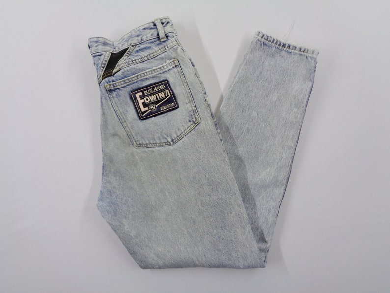 May include: Light blue acid wash denim jeans with a straight leg cut. The jeans feature a back pocket with a black and blue Edwin patch. The jeans are laid flat on a white surface.