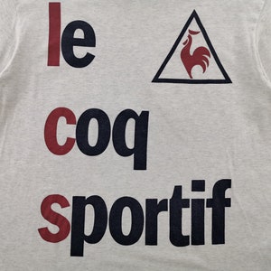 May include: Pale grey t-shirt featuring the text "le coq sportif" in navy and burgundy. A navy triangle with a burgundy rooster emblem is positioned above the word "sportif".