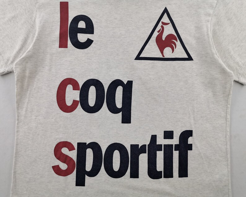 May include: Pale grey t-shirt featuring the text "le coq sportif" in navy and burgundy. A navy triangle with a burgundy rooster emblem is positioned above the word "sportif".