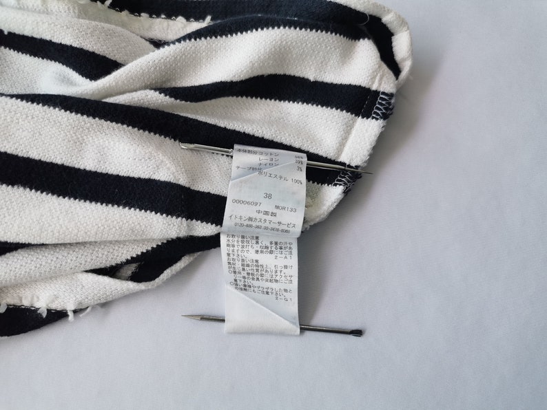 Puede incluir: Primer plano de una tela a rayas blancas y azul marino. Una etiqueta con texto japon&eacute;s y el n&uacute;mero 38 est&aacute; sujeta con alfileres. La tela parece ser un material de punto o tejido.