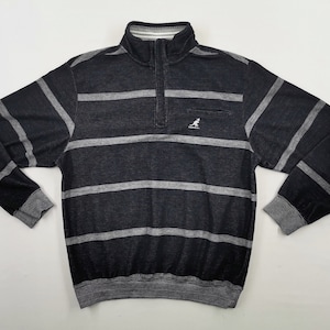May include: A black and gray striped sweatshirt with a quarter-zip collar and a small pocket on the chest. The sweatshirt has a ribbed collar, cuffs, and hem. The Kangol logo is visible on the pocket.