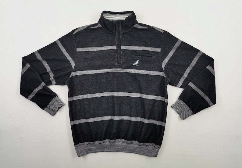 May include: A black and gray striped sweatshirt with a quarter-zip collar and a small pocket on the chest. The sweatshirt has a ribbed collar, cuffs, and hem. The Kangol logo is visible on the pocket.