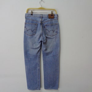 May include: Light blue denim jeans hanging on a yellow hanger. The jeans have a classic five-pocket design, a brown leather patch, and a red tag on the back pocket. The jeans are a light wash and appear to be new.