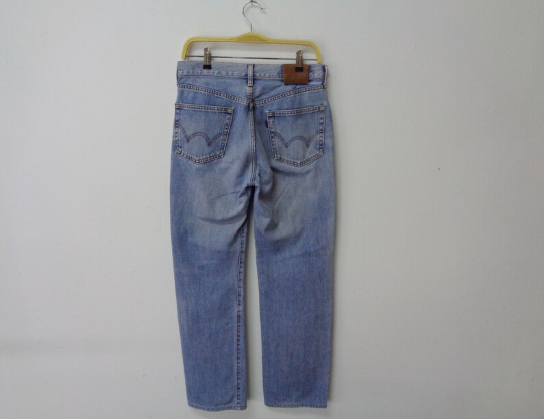 May include: Light blue denim jeans hanging on a yellow hanger. The jeans have a classic five-pocket design, a brown leather patch, and a red tag on the back pocket. The jeans are a light wash and appear to be new.