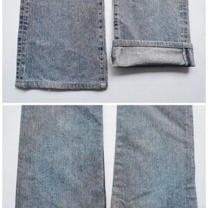 May include: Close-up of light blue denim jeans. The jeans have a faded wash and a slight flare at the bottom. One leg is cuffed, revealing a dark blue interior with red stitching. The other leg has a frayed hem.