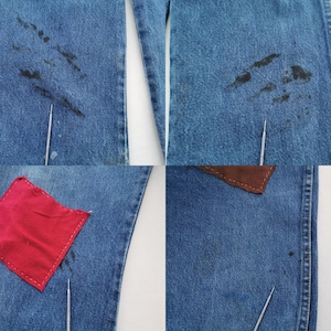 May include: Blue denim jeans featuring dark black stains on the upper thigh areas, a square red fabric patch stitched onto the lower leg, and a brown leather-like patch near the waist, with thin metal needles pointing to specific spots.