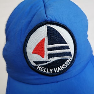 May include: A blue nylon cap with a circular patch featuring a sailboat design. The patch has a white background with a navy blue sailboat and a red sail. The text "HELLY HANSEN" is embroidered below the sailboat.