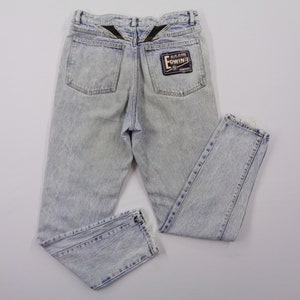 May include: Light blue acid wash jeans with a straight leg cut. The jeans feature two back pockets and a black patch with white text. The back of the waistband has a black design.