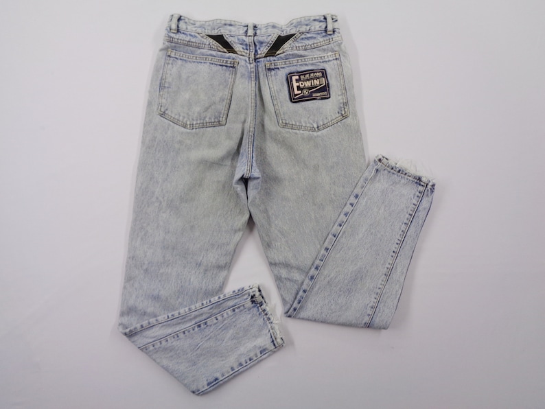 May include: Light blue acid wash jeans with a straight leg cut. The jeans feature two back pockets and a black patch with white text. The back of the waistband has a black design.