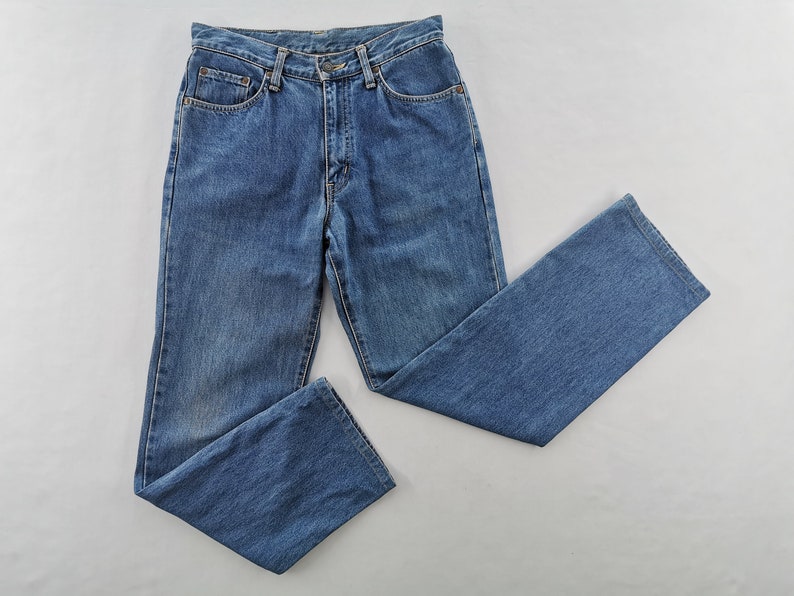 Puede incluir: Un par de vaqueros de mezclilla azules, con un dise&ntilde;o cl&aacute;sico de cinco bolsillos y corte recto. Los vaqueros est&aacute;n hechos de una tela de mezclilla de lavado medio y tienen cierre de bot&oacute;n y cremallera. Los vaqueros est&aacute;n planos sobre una superficie blanca.