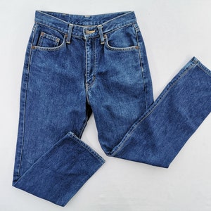 Puede incluir: Un par de vaqueros cl&aacute;sicos de color azul. Los vaqueros tienen una cintura alta, cierre de bot&oacute;n y cremallera, y cinco bolsillos. El denim es de un lavado medio y los vaqueros tienen una pernera recta. Los vaqueros est&aacute;n planos sobre una superficie blanca.