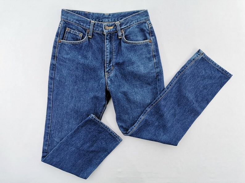 Puede incluir: Un par de vaqueros cl&aacute;sicos de color azul. Los vaqueros tienen una cintura alta, cierre de bot&oacute;n y cremallera, y cinco bolsillos. El denim es de un lavado medio y los vaqueros tienen una pernera recta. Los vaqueros est&aacute;n planos sobre una superficie blanca.