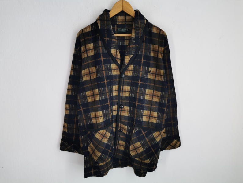 Puede incluir: Chaqueta vintage con botones y estampado de cuadros en azul marino, marr&oacute;n y beige. La chaqueta tiene cuello chal, dos bolsillos delanteros y un peque&ntilde;o logotipo en el pecho. Cuelga de una percha de madera.