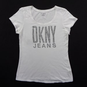 May include: White scoop-neck t-shirt featuring the DKNY JEANS logo in a glittery, textured font. The short-sleeved top is made of a soft material, suitable for casual wear.
