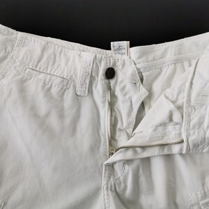 May include: Close-up of off-white cargo shorts with a button and zip closure. The shorts have pockets and visible stitching details. The fabric appears to be a sturdy cotton blend.