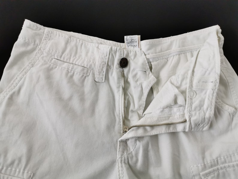 May include: Close-up of off-white cargo shorts with a button and zip closure. The shorts have pockets and visible stitching details. The fabric appears to be a sturdy cotton blend.