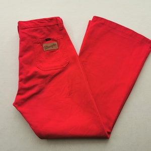 May include: Bright red Wrangler pants with a flared leg design. The pants feature a back pocket and a brown leather patch with the Wrangler logo. The pants are folded neatly, showcasing the fabric's texture and color.