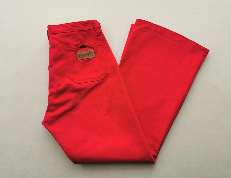 May include: Bright red Wrangler pants with a flared leg design. The pants feature a back pocket and a brown leather patch with the Wrangler logo. The pants are folded neatly, showcasing the fabric's texture and color.