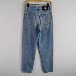 May include: Light blue denim jeans with a classic five-pocket design. The jeans feature a faded wash and a leather patch with a logo on the back. They are hanging on a wooden hanger.