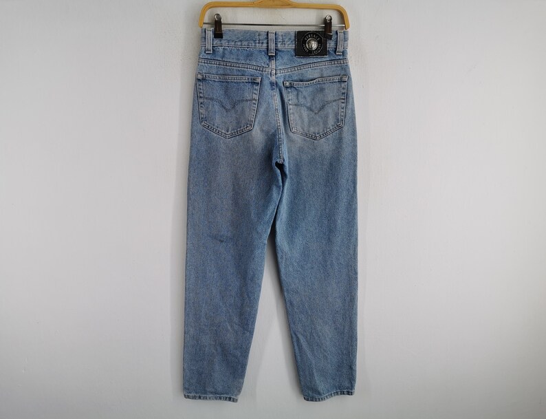 May include: Light blue denim jeans with a classic five-pocket design. The jeans feature a faded wash and a leather patch with a logo on the back. They are hanging on a wooden hanger.