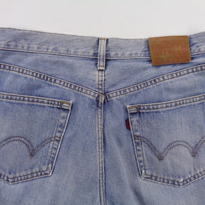 May include: Light blue denim jeans, rear view, showing the back pockets and the leather patch. The jeans have a classic five-pocket design with a red tab on the right pocket. The stitching is a contrasting brown color.