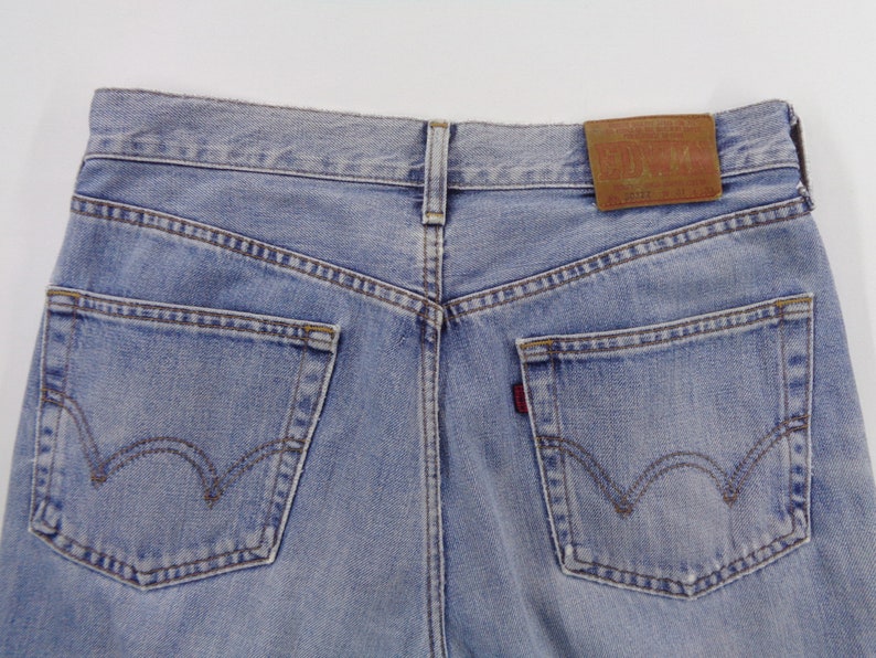 May include: Light blue denim jeans, rear view, showing the back pockets and the leather patch. The jeans have a classic five-pocket design with a red tab on the right pocket. The stitching is a contrasting brown color.