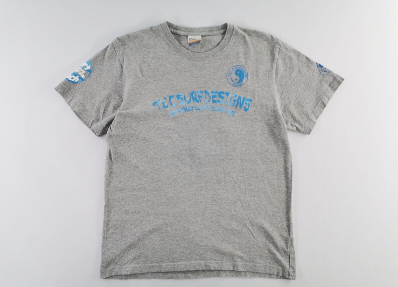 May include: A heather gray t-shirt with short sleeves. The shirt features the text "TEC SURF DESIGNS" in blue, along with a yin-yang symbol and additional text on the sleeves. A casual, everyday wear tee.