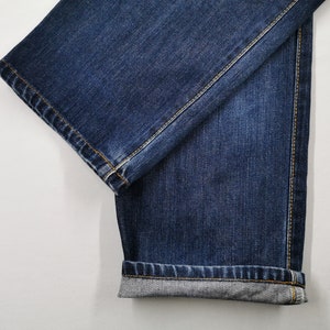 May include: Close-up of dark blue denim jeans. The jeans are folded, showing the hem and the stitching. The fabric has a textured appearance, and the seams are highlighted with yellow thread. The jeans are on a white background.