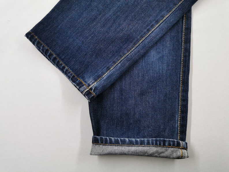 May include: Close-up of dark blue denim jeans. The jeans are folded, showing the hem and the stitching. The fabric has a textured appearance, and the seams are highlighted with yellow thread. The jeans are on a white background.
