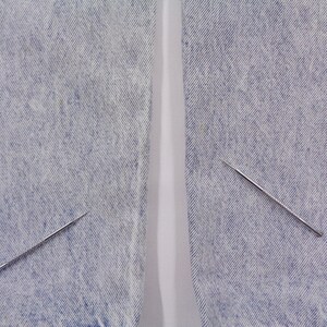 May include: Two silver sewing needles with textured shafts are positioned on a light blue denim fabric. The needles are angled, with one near the bottom left and the other near the top right. The fabric has a faded appearance.