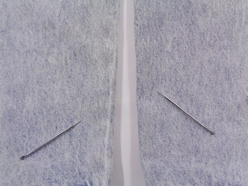 May include: Two silver sewing needles with textured shafts are positioned on a light blue denim fabric. The needles are angled, with one near the bottom left and the other near the top right. The fabric has a faded appearance.