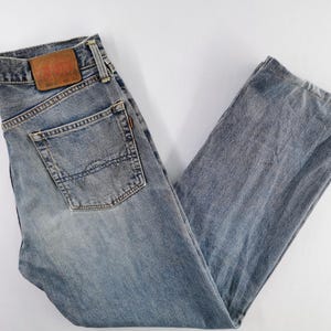 Puede incluir: Vaqueros de mezclilla azul claro con un dise&ntilde;o cl&aacute;sico de cinco bolsillos. Los vaqueros presentan un lavado descolorido y un parche de cuero marr&oacute;n en la parte trasera. Los vaqueros est&aacute;n planos sobre una superficie blanca.