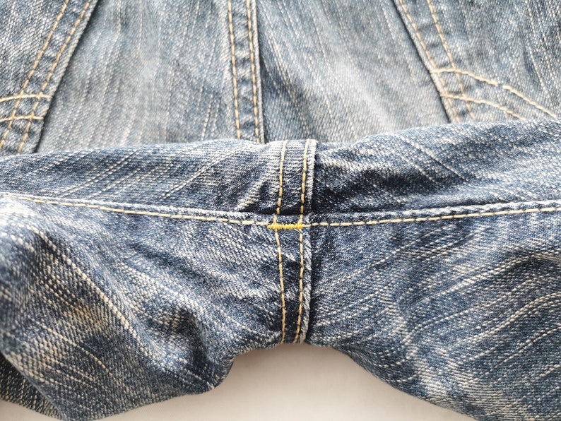 May include: Close-up of blue denim jeans, showcasing the texture and stitching details. The image highlights the seams and the fabric's weave, with yellow thread used for the stitching.