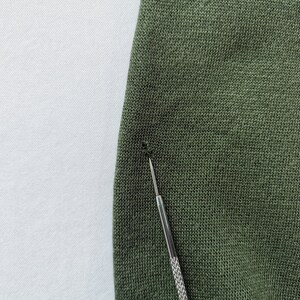 May include: Close-up of olive green knit fabric with a small hole. A silver tool is inserted into the fabric, highlighting the damage. The fabric has a textured appearance.