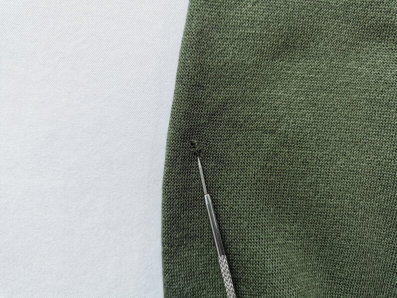 May include: Close-up of olive green knit fabric with a small hole. A silver tool is inserted into the fabric, highlighting the damage. The fabric has a textured appearance.