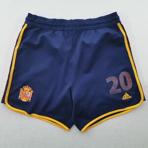 May include: Navy blue athletic shorts featuring a yellow trim along the edges, an elastic waistband, and the number 20 printed in a textured grey font. The shorts display the Spanish national coat of arms and a small yellow Adidas logo on the front.