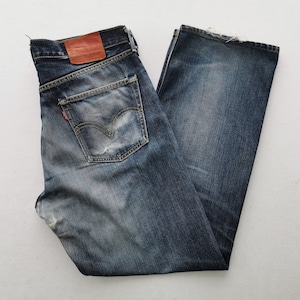 May include: A pair of dark blue denim jeans featuring a faded wash, a tan leather patch on the back waistband, a single back pocket with a red tab, and frayed edges at the hem. The denim displays natural wear patterns and whiskering throughout.