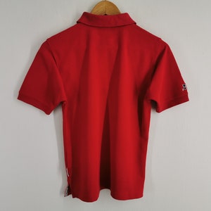 K&ouml;nnte beinhalten: Ein leuchtend rotes Poloshirt mit kurzen &Auml;rmeln, Kragen und einer Knopfleiste mit zwei Kn&ouml;pfen. Das Shirt hat ein kleines Logo auf dem linken &Auml;rmel und einen wei&szlig;en Streifen an der Seite. Es h&auml;ngt an einem Holzkleiderb&uuml;gel.
