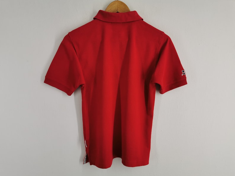 K&ouml;nnte beinhalten: Ein leuchtend rotes Poloshirt mit kurzen &Auml;rmeln, Kragen und einer Knopfleiste mit zwei Kn&ouml;pfen. Das Shirt hat ein kleines Logo auf dem linken &Auml;rmel und einen wei&szlig;en Streifen an der Seite. Es h&auml;ngt an einem Holzkleiderb&uuml;gel.
