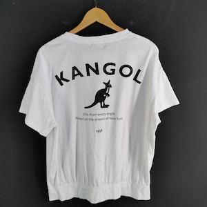 Pu&ograve; includere: T-shirt bianca con la scritta nera "KANGOL" e un logo a forma di canguro sul retro. Sotto il logo, il testo recita "Chic from every angle. Raised on the streets of New York. 1938."