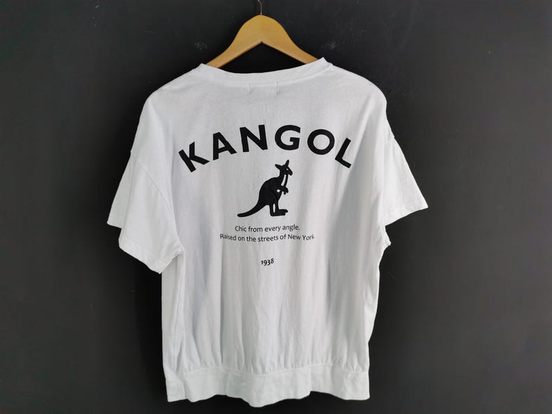 Pu&ograve; includere: T-shirt bianca con la scritta nera "KANGOL" e un logo a forma di canguro sul retro. Sotto il logo, il testo recita "Chic from every angle. Raised on the streets of New York. 1938."