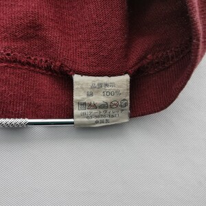 May include: Close-up of a burgundy fabric with a sewn seam. A rectangular tag is attached, displaying Japanese text and symbols, including "100%" and care instructions. The tag is white with black text.